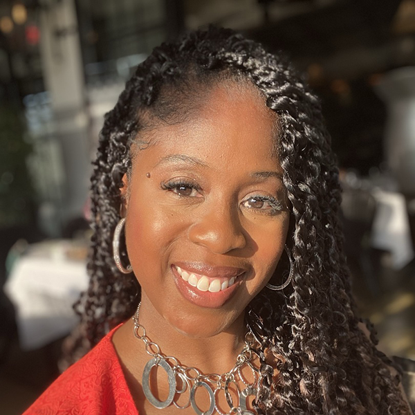 Smiling woman with curly hair, wearing earrings and a necklace, with sunlight on her face.