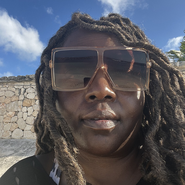 Close-up of a woman wearing large sunglasses with a stone wall in the background.